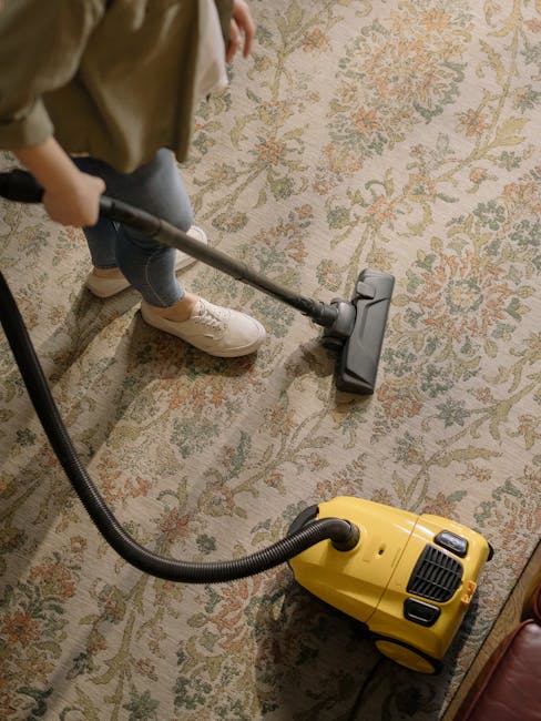 A person kneeling on a patterned area rug in a domestic setting, preparing a portable vacuum cleaner with a yellow body and black hose for surface cleaning. The individual is wearing a beige jacket and blue jeans, and is opening the vacuum's compartment to access attachments. The room features warm lighting, and the carpet shows a detailed, ornate pattern with neutral tones. This scene reflects routine maintenance and deep cleaning efforts, consistent with residential surface cleaning and sanitisation services provided by Acton Carpet Cleaning, as highlighted on their website's page about quick guides for carpet cleaning near Acton Town station.