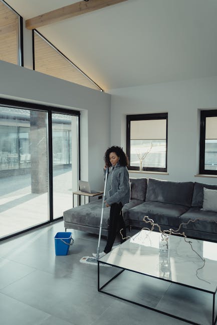 A woman with curly hair is engaged in surface cleaning within a modern living room, using a mop on a large, light gray tiled floor that appears clean and polished. She is wearing a gray top and black pants, holding the mop with a mop handle visible, and standing near a blue cleaning bucket placed on the floor. The room features a gray upholstered sectional sofa positioned against the wall, with two black-framed windows behind it, allowing natural light to illuminate the space. There is a glass coffee table with a slim black metal frame in the foreground, and a sliding glass door on the left side reveals an outdoor patio area. The room has white walls and a ceiling with wooden beams, creating a bright, airy atmosphere suitable for detailed surface cleaning and maintenance, consistent with the services offered by Acton Carpet Cleaning regarding end of tenancy cleaning and sanitisation.