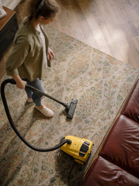 A person kneeling on a patterned carpet in a domestic setting, opening a yellow and black vacuum cleaner with a flexible hose. The carpet features intricate floral designs in muted tones, and the room appears well-lit with natural or warm lighting. The individual is wearing casual jeans and a beige top or jacket, focused on preparing the vacuum for deep cleaning or maintenance. The scene highlights surface cleaning and routine maintenance as part of domestic cleaning services offered by Acton Carpet Cleaning, emphasizing hygiene and carpet care.