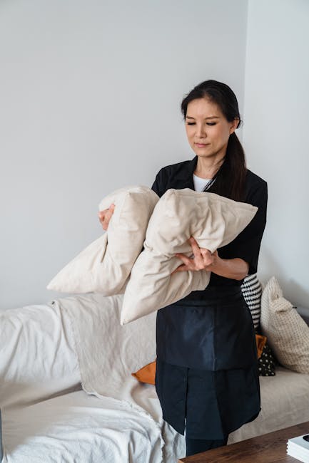 A woman in a black uniform holding two beige cushions in a bright, neatly maintained living room with white walls and a light-colored sofa. The room features a wooden table, visible cushions, and natural lighting. The scene reflects domestic cleaning and upholstery refreshing by Acton Carpet Cleaning, emphasizing surface cleaning and tidiness in a home setting.
