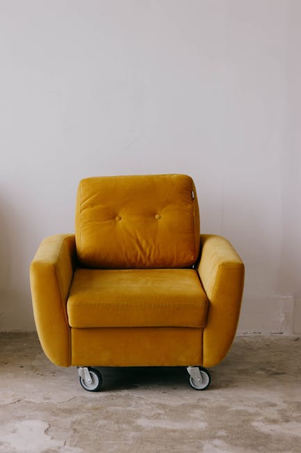 A person wearing a black jacket is using a handheld vacuum cleaner to deep clean a decorative yellow and white patterned cushion on a dark fabric sofa in a residential living room. The sofa's surface appears clean and dust-free, with a smooth and well-maintained textile finish. The room is well-lit, highlighting the sleekness of the black sofa and the vibrant design of the cushion. The vacuum cleaner attachment is positioned over the cushion, emphasizing surface cleaning and maintenance procedures, with no other objects visible in the immediate area. This image illustrates upholstery cleaning as part of routine domestic surface care, as provided by Acton Carpet Cleaning.